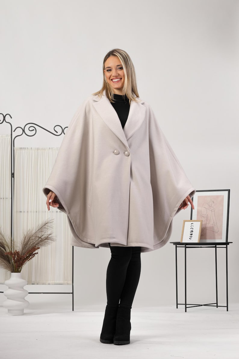 May include: A light beige cape coat with a double-breasted closure and two large buttons. The coat has a wide, flowing design and is worn over a black top and leggings. The model is smiling.