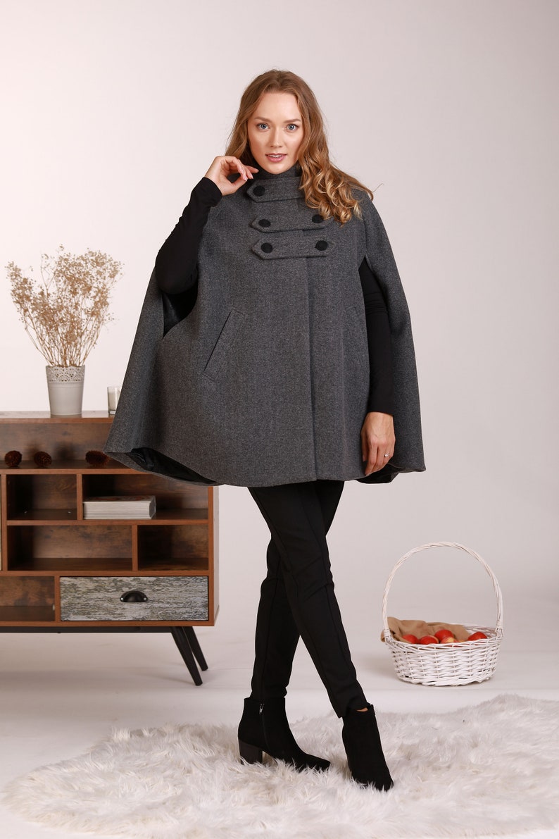 May include: A gray wool cape with a high collar and four black buttons. The cape has a fitted silhouette and is worn with black pants and black heeled boots.