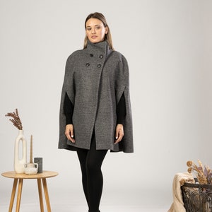 May include: A charcoal gray wool-blend cape coat featuring a high collar, a double-breasted front with four black buttons, and arm slits. The garment is worn over a black long-sleeved top, black leggings, and black ankle boots.