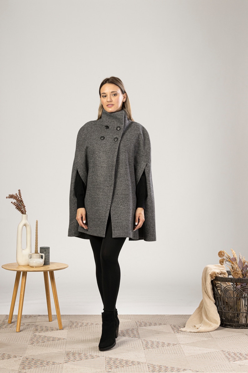 May include: A charcoal gray wool-blend cape coat featuring a high collar, a double-breasted front with four black buttons, and arm slits. The garment is worn over a black long-sleeved top, black leggings, and black ankle boots.