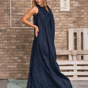 K&ouml;nnte beinhalten: Ein &auml;rmelloses, bodenlanges marineblaues Kleid. Das Kleid hat ein schlichtes, elegantes Design mit einem flie&szlig;enden Rock. Das Model steht vor einer Backsteinmauer und einem hellgr&uuml;nen Stoff.
