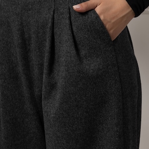 May include: Close-up of charcoal gray pleated trousers with a hand in the pocket. The pants have a high waist and a tailored fit. The fabric appears to be a soft wool blend, suitable for cooler weather.