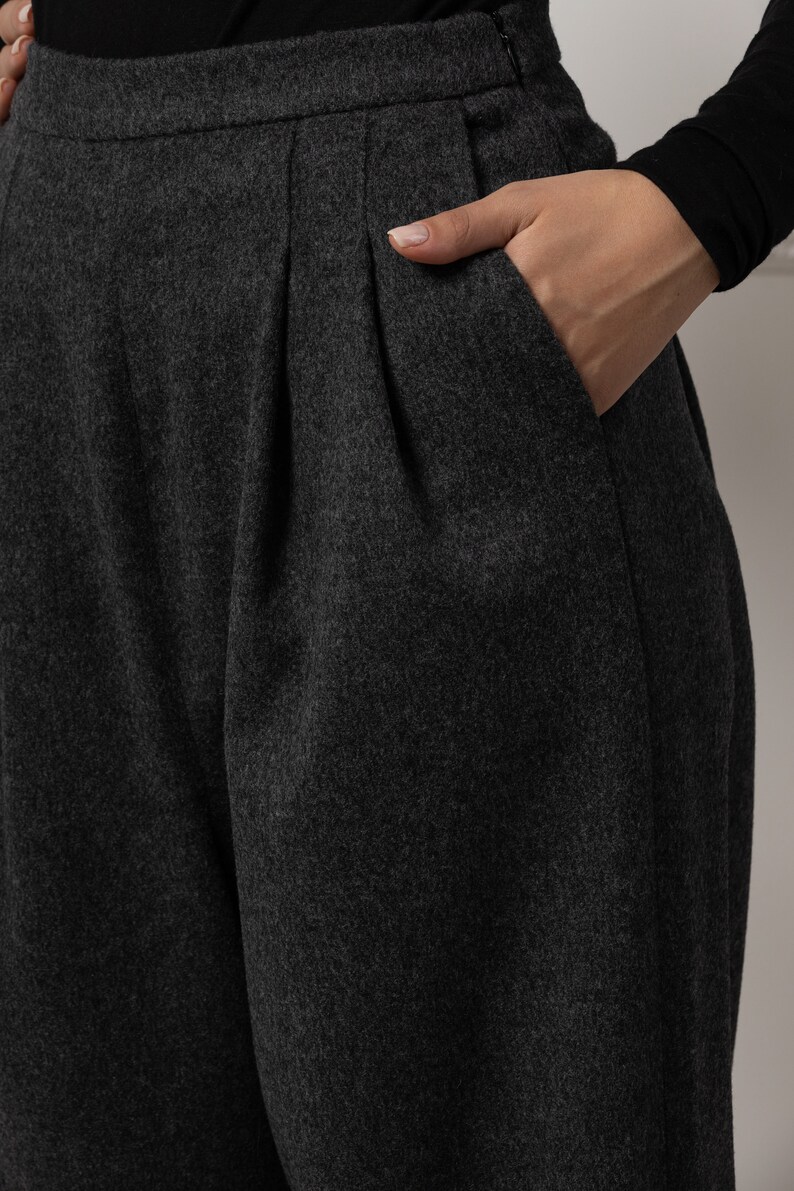 May include: Close-up of charcoal gray pleated trousers with a hand in the pocket. The pants have a high waist and a tailored fit. The fabric appears to be a soft wool blend, suitable for cooler weather.