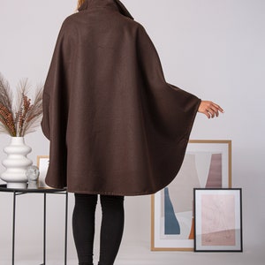 May include: A brown cape with a high collar. The cape is made of a soft material and has a flowing design. The model is wearing black leggings and black heeled boots. The background includes framed artwork and decorative items.