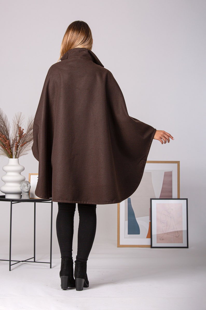 May include: A brown cape with a high collar. The cape is made of a soft material and has a flowing design. The model is wearing black leggings and black heeled boots. The background includes framed artwork and decorative items.