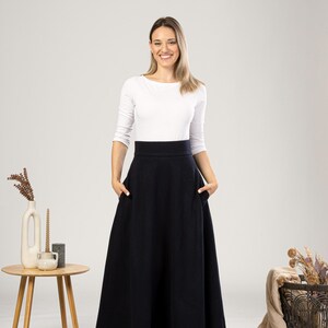 May include: A woman wearing a white long-sleeve top and a long, dark navy blue skirt with pockets. The skirt has a high waist and a full, flowing silhouette. The woman is smiling, and the background is a neutral color.