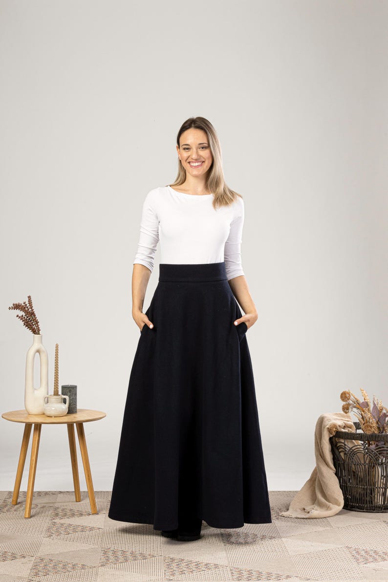 May include: A woman wearing a white long-sleeve top and a long, dark navy blue skirt with pockets. The skirt has a high waist and a full, flowing silhouette. The woman is smiling, and the background is a neutral color.
