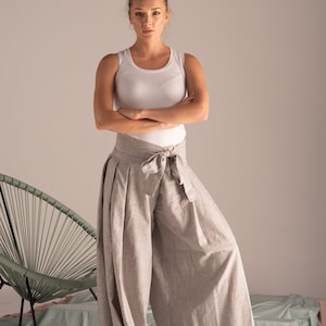 May include: A woman in a white vest top and light grey wide-leg trousers with a matching tie at the waist. The trousers have a pleated design. The background includes a green chair and a light blue sheet.