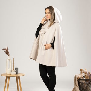 May include: A beige hooded cape with a button closure and a pocket. The cape is worn by a person with black leggings and black ankle boots.