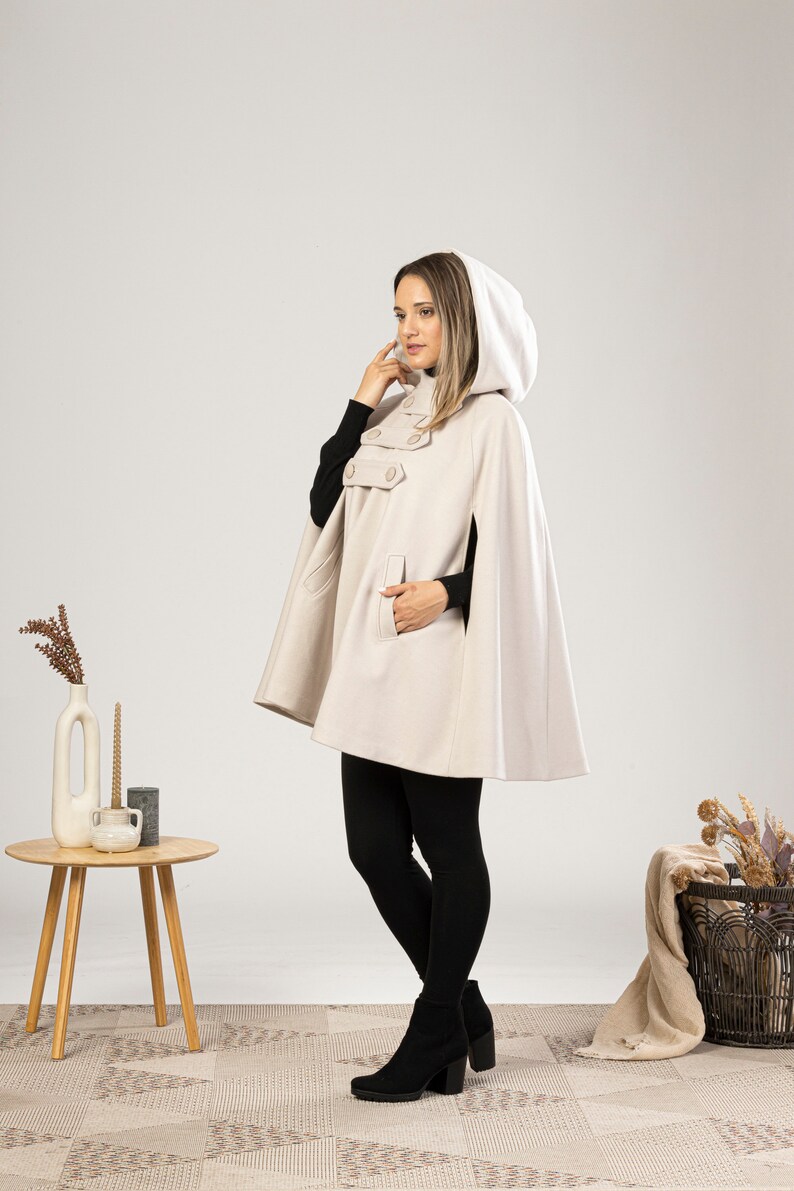 May include: A beige hooded cape with a button closure and a pocket. The cape is worn by a person with black leggings and black ankle boots.