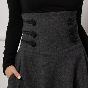 May include: A charcoal grey skirt with a high-waisted design and a flared silhouette. The skirt features decorative black leather straps and buttons on the waist. The model is wearing a black long-sleeved top.