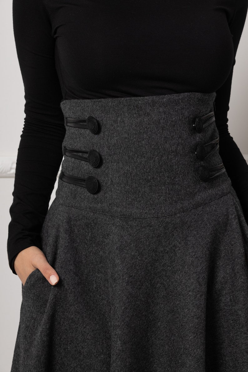 May include: A charcoal grey skirt with a high-waisted design and a flared silhouette. The skirt features decorative black leather straps and buttons on the waist. The model is wearing a black long-sleeved top.