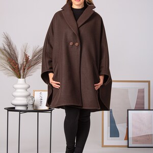 May include: A brown cape coat with a wide collar and two large buttons. The coat has a relaxed fit and is worn over a black top and leggings. The model is wearing black ankle boots. The coat is a stylish outerwear option.