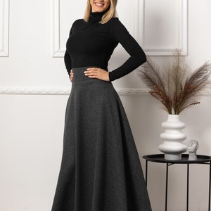 May include: A woman wearing a black turtleneck long-sleeve shirt and a gray, flared maxi skirt. The skirt has a high waist and falls to the ankles. The woman is smiling and posing in front of a white wall.