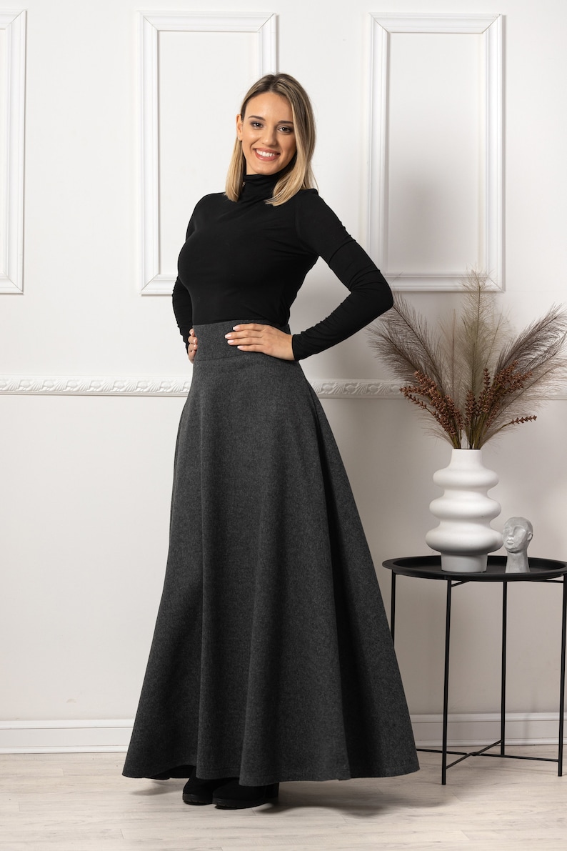 May include: A woman wearing a black turtleneck long-sleeve shirt and a gray, flared maxi skirt. The skirt has a high waist and falls to the ankles. The woman is smiling and posing in front of a white wall.