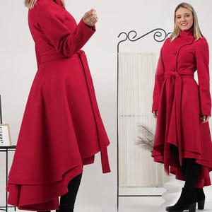 May include: A vibrant red long-sleeved coat featuring a high-low hemline, a structured collar, and a matching fabric tie belt at the waist. The coat has a button-front closure and a flared, layered skirt design that falls below the knees.