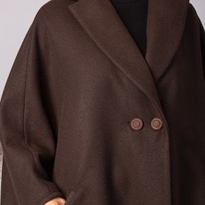 May include: A dark brown wool coat with a wide lapel collar and two large, round buttons. The coat has a double-breasted design and a relaxed fit. The model is wearing a black turtleneck underneath.