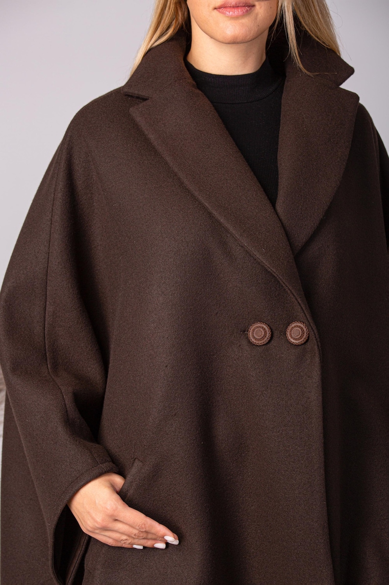 May include: A dark brown wool coat with a wide lapel collar and two large, round buttons. The coat has a double-breasted design and a relaxed fit. The model is wearing a black turtleneck underneath.