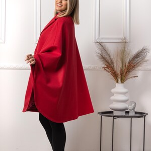 May include: A vibrant red cape coat with a wide collar. The coat is worn over black leggings and black heeled boots. The woman is smiling. The background is a white wall with decorative frames.