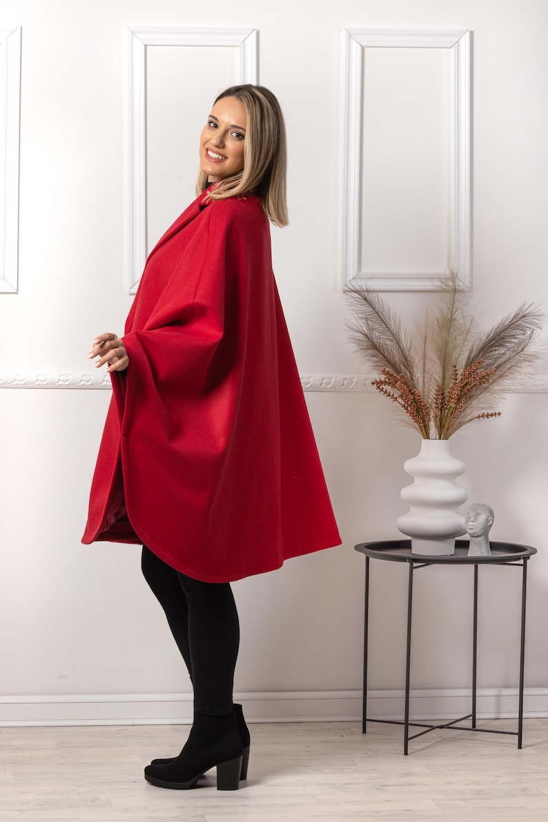 May include: A vibrant red cape coat with a wide collar. The coat is worn over black leggings and black heeled boots. The woman is smiling. The background is a white wall with decorative frames.