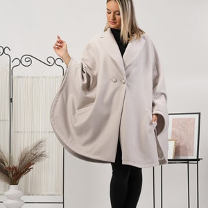 May include: A light beige cape coat with a classic collar and two large buttons. The coat has a unique cape-like design with wide sleeves and pockets. The model is wearing black pants and boots.