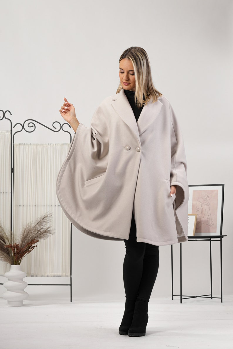 May include: A light beige cape coat with a classic collar and two large buttons. The coat has a unique cape-like design with wide sleeves and pockets. The model is wearing black pants and boots.