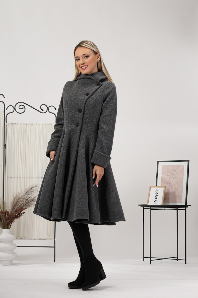 May include: A charcoal grey wool-blend coat featuring a high collar, long sleeves with folded cuffs, and a flared, pleated skirt silhouette. The coat has a diagonal button closure with three visible black buttons and side pockets.