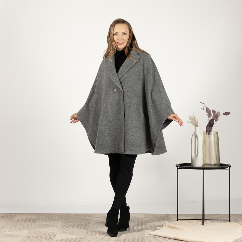 May include: A gray cape coat with a button closure and a notched collar. The coat is worn over a black turtleneck and black pants. The model is wearing black boots. The background is white.