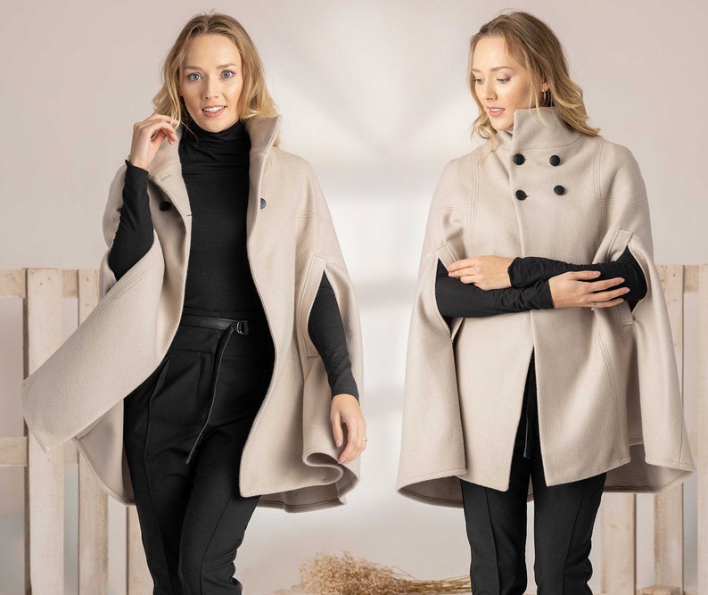 May include: A beige wool-blend cape coat featuring a high collar, structured shoulders, and a double-breasted front with four large black circular buttons. The garment has arm slits on the sides and a flared hemline, worn over a black turtleneck.