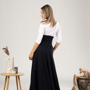 May include: A navy blue maxi skirt with a high waist, worn with a white long-sleeved top. The skirt has a flared design. The model is viewed from the back, highlighting the skirt's shape and style. The skirt's length is approximately 100 cm.
