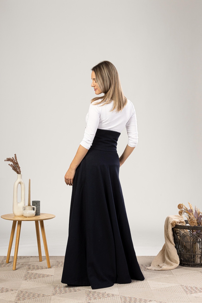 May include: A navy blue maxi skirt with a high waist, worn with a white long-sleeved top. The skirt has a flared design. The model is viewed from the back, highlighting the skirt's shape and style. The skirt's length is approximately 100 cm.