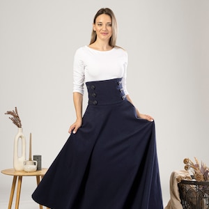 May include: A navy blue maxi skirt with a high-waisted design and button detailing. The skirt is worn with a white long-sleeved top. The model is standing in front of a neutral background.
