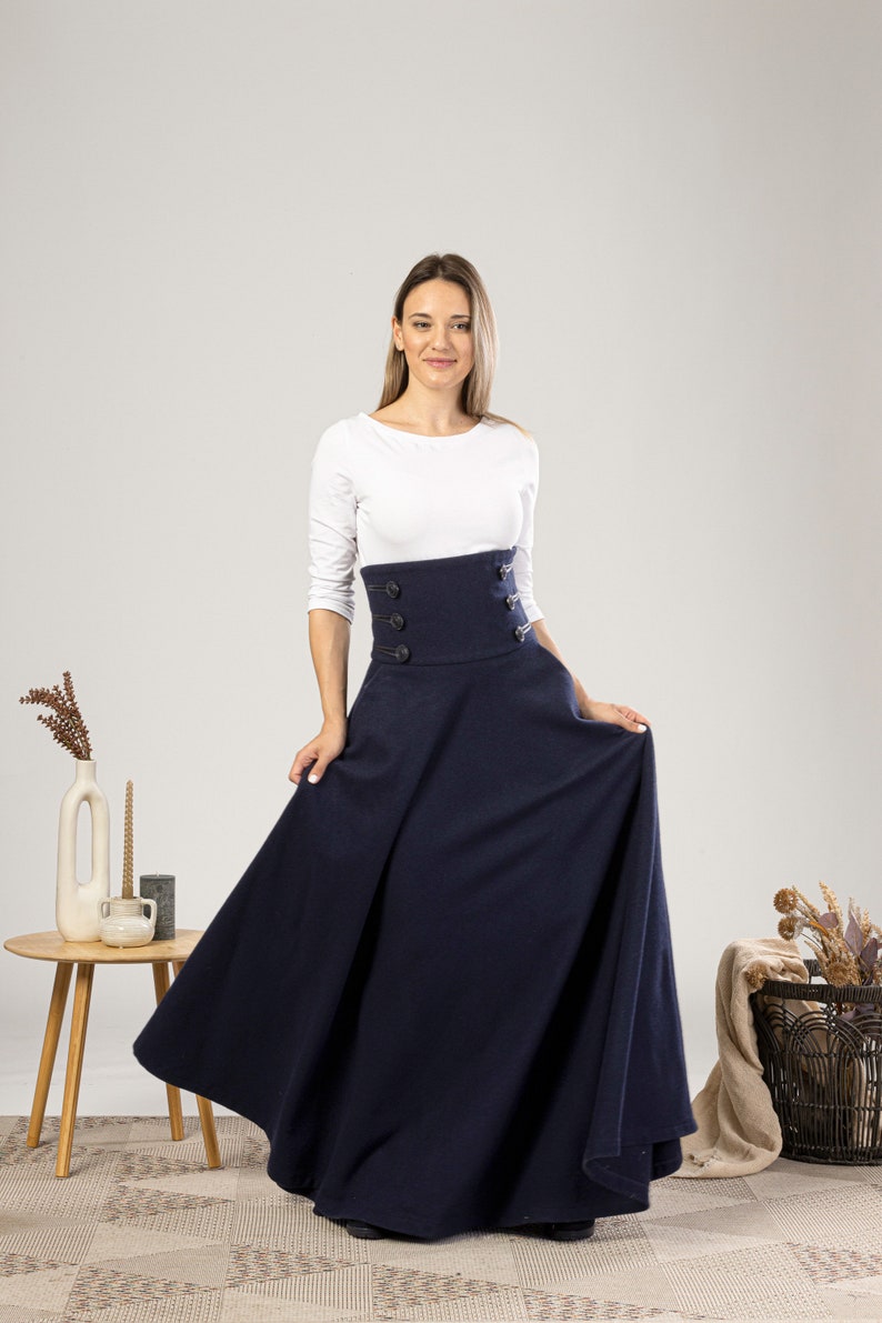 May include: A navy blue maxi skirt with a high-waisted design and button detailing. The skirt is worn with a white long-sleeved top. The model is standing in front of a neutral background.
