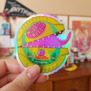 May include: A holographic sticker of a yellow and green Hubba Bubba bubble gum container with a pink and purple gum inside. The container is open and has teeth. The text on the sticker reads "Hubble Bubble,  Toil and Trouble Gum,  SourApple, 97% Poison Free".