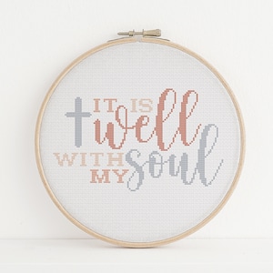 Puede incluir: Un aro de bordado blanco con la frase "It is well with my soul" bordada en hilo gris, rosa y melocotón. Una cruz está bordada en hilo gris sobre la frase.