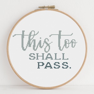 Puede incluir: Un aro de bordado blanco con la frase "This too shall pass" bordada en hilo gris.