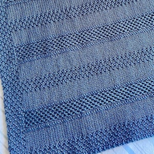 May include: A blue knitted blanket with a textured pattern. The blanket is made with a fine yarn and has a soft, cozy feel.