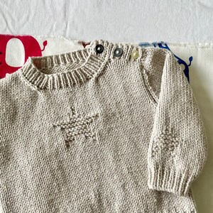 May include: A beige knitted baby sweater with a star design on the front and button closures on the shoulder.