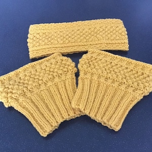 May include: A set of three knitted accessories in a light yellow color. The set includes a headband and two leg warmers. The headband and leg warmers have a textured, raised pattern. The leg warmers are approximately 6 inches long.
