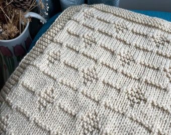May include: A cream-colored knitted blanket with a textured pattern. The blanket is folded and placed on a blue and white surface.