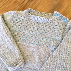 Peut inclure: Un pull en tricot gris avec un motif géométrique bleu et vert sur le devant. Le pull a un col rond et des manches longues. Il y a trois boutons bleus sur l'épaule droite.