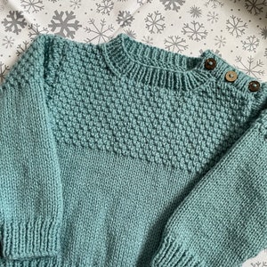 Könnte beinhalten: Ein tealblauer Strickpullover mit einem strukturierten Muster und Knopfverschlüssen an der Schulter.