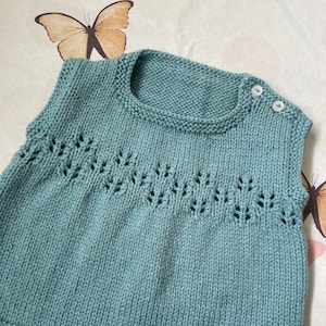May include: A teal blue knitted sleeveless vest with a delicate lace pattern around the neckline. The vest has a round neckline and two white buttons on the shoulder.