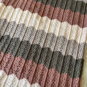 Può includere: Una copertina per neonati lavorata a maglia con un motivo a chevron in tonalità di bianco, grigio e rosa.