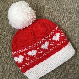 以下が含まれることがあります： 赤と白のニット帽。上部に大きな白いポンポンが付いています。赤いクラウンには白いハート模様があり、つばは白です。寒い季節にぴったりの冬用帽子です。