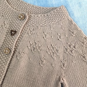 May include: A beige knitted cardigan with a button fastening and a delicate stitch pattern. The cardigan has a relaxed fit and a slightly cropped length.
