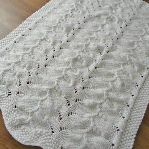 May include: White knitted baby blanket with a delicate leaf pattern. The blanket is made with a soft yarn and has a crocheted border.