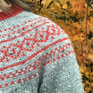 May include: A grey knitted jumper with a red and white geometric pattern. The jumper is made with a chunky yarn and has a relaxed fit.