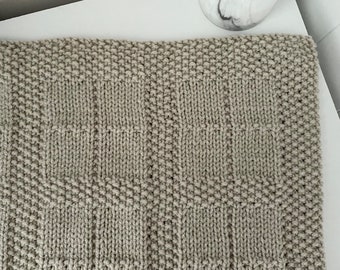 May include: A beige knitted throw blanket with a geometric pattern. The blanket is made with a textured yarn and has a subtle sheen.