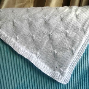 May include: A white knitted baby blanket with a delicate diamond pattern. The blanket is soft and fluffy, perfect for cuddling a newborn.